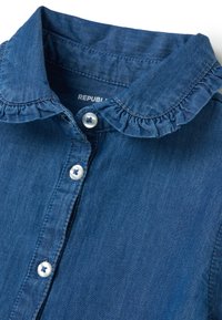 Camicia di jeans con colletto arricciato, tessuto azzurro chiaro, chiusura frontale con bottoni decorativi blu e bianchi.