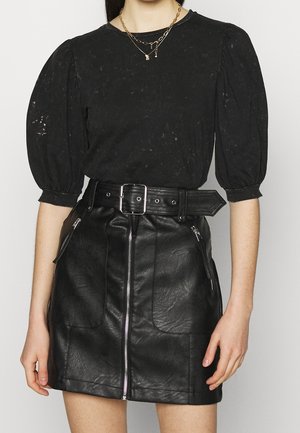 Kobieta w czarnej bluzce z bufkami oraz czarnej mini spódnicy z przodu na zamek i szerokim paskiem z srebrną klamrą.