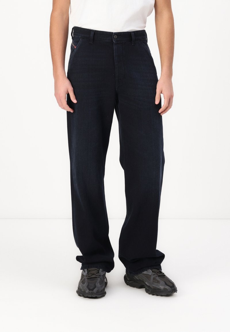 Personne portant un jean bleu foncé ample, une chemise blanche et des baskets noires, debout sur un fond blanc uni. Seule la moitié inférieure est visible.