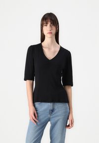 Sort kortærmet top med V-halsdesign, indsyet talje og blød tekstur. Parret med lyse jeans, modellen er placeret mod en neutral baggrund.