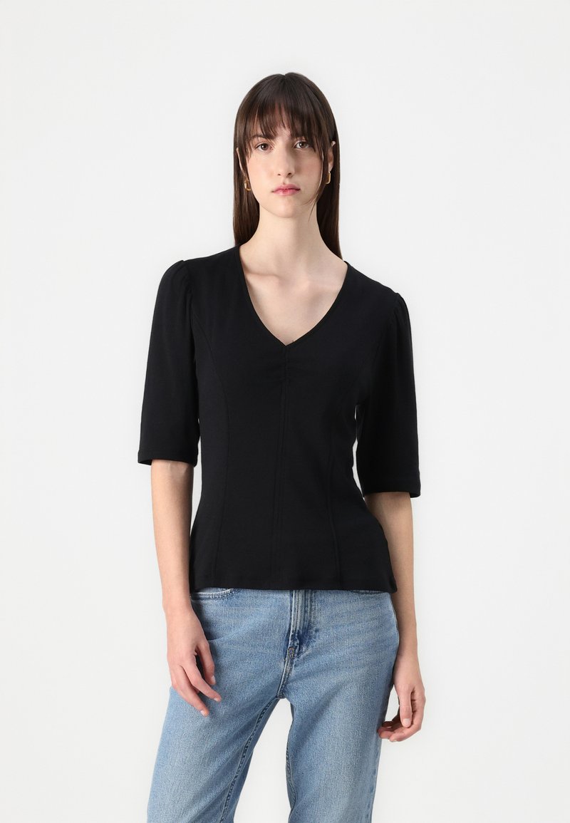 Sort kortærmet top med V-halsdesign, indsyet talje og blød tekstur. Parret med lyse jeans, modellen er placeret mod en neutral baggrund.