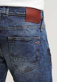 Jeans in denim di blu chiaro e scuro con una texture usurata. Le caratteristiche includono una toppa in pelle sulla cintura etichettata "LTB" e cuciture a contrasto.