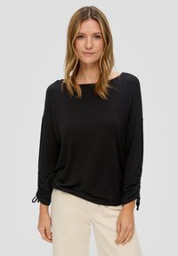 Schwarzes, locker sitzendes Longsleeve mit weitem Ausschnitt, eingeknöpften Ärmeln mit Bindebändern und weichem, glattem Stoff. Kombiniert mit hellen Hosen.