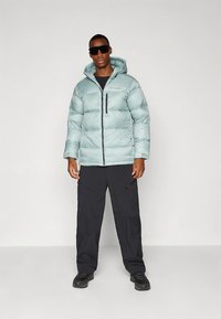 Lichtblauwe donsjas met capuchon, voorzien van een ritssluiting aan de voorkant en een zak; gecombineerd met zwarte cargobroeken en laarzen. Het oppervlak is glad.