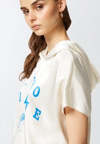 Felpa con cappuccio da donna in satin crema con lettering grafico blu. Presenta una vestibilità comoda e maniche a taglio vivo per un look casual.