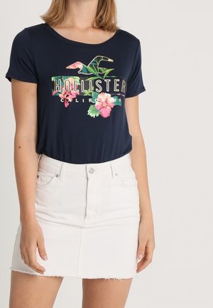 Granatowy t-shirt z krótkim rękawem z kwiatowym nadrukiem i napisem "HOLLISTER CALIFORNIA", zestawiony z białą mini spódnicą jeansową z postrzępionym dołem.