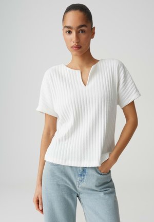 Femme aux cheveux tirés en arrière portant un t-shirt blanc à manches courtes côtelé et un jean bleu clair, debout avec une main dans la poche.