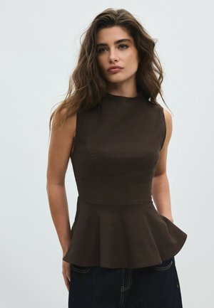 Femme aux longs cheveux ondulés porte un top péplum marron foncé sans manches et un pantalon foncé, debout devant un fond clair uni.