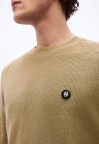 Άνδρας που φοράει μπεζ υφασμένο πουλόβερ με μικρό μαύρο κυκλικό έμβλημα στο στήθος που απεικονίζει ένα στυ λιζαρισμένο λογότυπο "B".