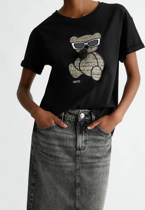 Personne portant un T-shirt noir avec un ours en peluche texturé portant des lunettes de soleil blanches et une jupe en jean grise, les mains dans les poches.