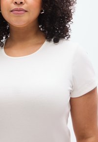 Άσπρο ριμπ μπλουζάκι με στρογγυλή λαιμόκοψη και κοντά μα νίκια, με διακριτική υφή. Το φόντο είναι απλό και διακριτικό.