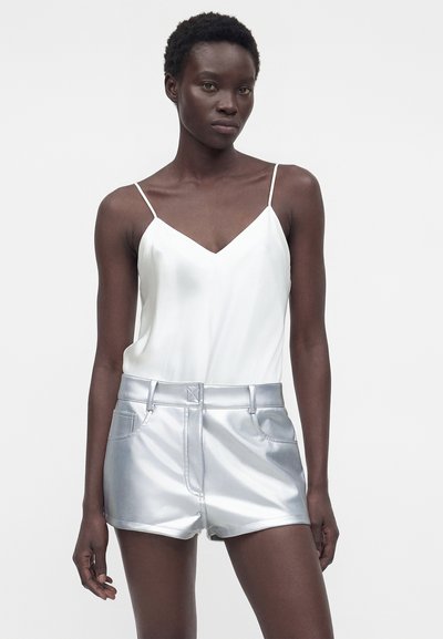 Femme aux cheveux courts portant un haut blanc sans manches et un short argenté brillant, debout devant un fond clair uni.