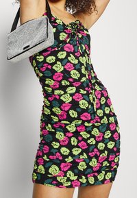 Schwarzes gerafftes Kleid mit bunten Blumenmustern in Pink, Grün und Blaugrün. Kombiniert mit einer kleinen silbernen Handtasche mit schwarzem Rand.