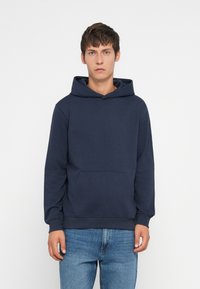 Granatowa bluza z kapturem z miękkiego materiału, z przednią kieszenią, sznurkiem do regulacji i ściągaczami na mankietach. Noszona z jasnoniebieskimi dżinsami.