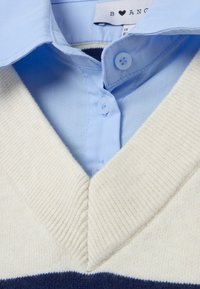 Maglione a V in crema con righe blu navy e texture a coste. Camicia azzurra con colletto sottostante, dotata di due bottoni azzurri.