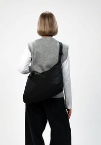 Černá taška přes rameno z lehké látky, s zakřiveným tvarem, nastavitelným popruhem a minimalistickým designem bez viditelných kovových prvků.