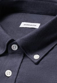 Camicia button-up blu scuro con bottoni bianchi, che mostra il colletto e un'etichetta bianca con testo del marchio all'interno della scollatura.