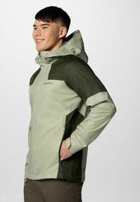 Uśmiechnięty młody mężczyzna w dwukolorowej zielonej kurtce Columbia z kapturem, stojący z jedną ręką w kieszeni, zwrócony w lewo na białym tle.