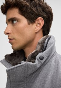 Graue Jacke mit einem Kunstfellkragen, ausgestattet mit einem Reißverschluss und großem Kragen. Hergestellt aus strukturiertem Stoff, mit einem schwarzen Knopfdetail.