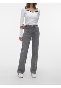 Femme portant un haut blanc à manches longues avec un bord noir, un jean gris taille haute à jambes larges, et des baskets blanches avec des accents rouges et bleus.