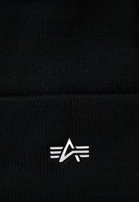 Gorro de punto negro con un logo plateado que presenta líneas estilizadas y un acento triangular. La textura es suave y está tejido de manera apretada.
