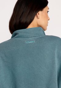 Teal pullover med høj krave, lavet af blødt stof; har broderet "VOLCOM" logo i lys blå på ryggen.