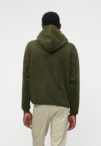 Σκούρος πράσινος φλις ζακέτα με κουκούλα και υφή, με χαλαρή εφαρμογή και μακριά μανίκια, συνδυασμένη με ανοιχτό μπεζ παντελόνι.