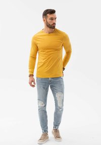 Langsleeve mosterdkleurig shirt, gecombineerd met versleten lichtblauwe jeans en beige sneakers, met een ontspannen pasvorm en een gladde textuur.
