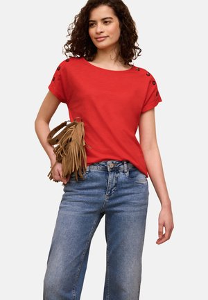 Vrouw die een rood kortärmelig shirt met knoopdetails draagt, blauwe spijkerbroek en een bruine fringed handtas vasthoudt tegen een witte achtergrond.