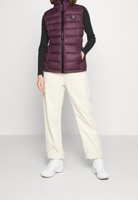 Fialová péřová vesta s vysokým límcem, zapínáním na zip a logem; nošená přes černé tri čko s dlouhým rukávem a doplněná krémovými kalhotami.