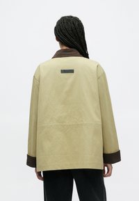 Beige jakke med brun fløjlsribskrave og manchetter, afslappet pasform, med et sort mærke på ryggen, lavet af tekstureret, letvægtsstof.