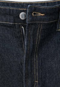 Pantalones vaqueros de mezclilla azul oscuro con botón plateado visible y cremallera parcialmente cerrada, con costuras color cuero alrededor de la cintura y el área de la bragueta.