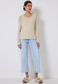 Strickpullover in Creme mit V-Ausschnitt, kombiniert mit hellblauen weit geschnittenen Jeans und braunen, flauschigen Slidesandalen. Verfügt über eine strukturierte Naht an den Schultern.