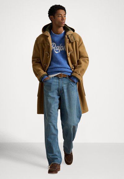 Καφέ μάλλινο παλτό τύπου νταφλ, μπλε πλεκτό πουλόβερ με την εκτύπωση «Ralph», ανοιχτό μπλε τζιν και καφέ μοκασίνια, με τα χέρια στις τσέπες.