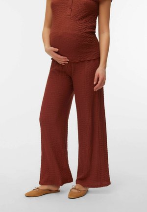 Nosečnica, ki nosi ohlapne hlače teksturirane rjave barve in ujeta zgornji del, z eno roko na trebuhu in tanjšimi ravnimi čevlji.