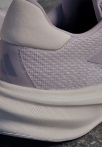 Zapatilla deportiva de color púrpura claro con parte superior de malla texturizada, cuello acolchado suave y suela de goma blanca lisa con surcos y costuras visibles.