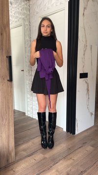 Mlada žena u crnoj majici bez rukava, plisiranoj suknji i sjajnim crnim čizmama do koljena, drži presavijenu ljubičastu jaknu u zatvorenom prostoru.