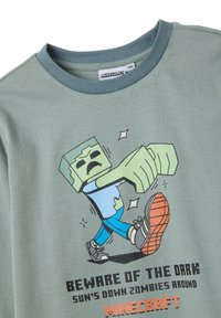 Lichtgroene katoenen t-shirt met een cartoonfiguur met een vierkante kop en uitgestrekte arm. Onder de afbeelding staat de tekst "Pas op voor het Donkere".