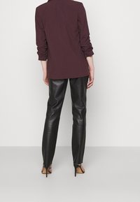 Henkilö seisoo selin päin pukeutuneena tummanpunainen bleiseri, mustat nahkahousut ja mustat korkeakorkoiset sandaalit valkoisella taustalla.