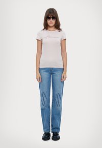 T-shirt aderente rosa chiaro con logo "Armani" in corsivo, abbinata a jeans a gamba dritta blu con un leggero motivo grafico e scarpe nere.