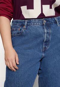 Pantalones vaqueros de mezclilla azul de tiro alto con un acabado texturizado, costuras naranjas y bolsillos delanteros. Combinados con una sudadera burdeos por encima.