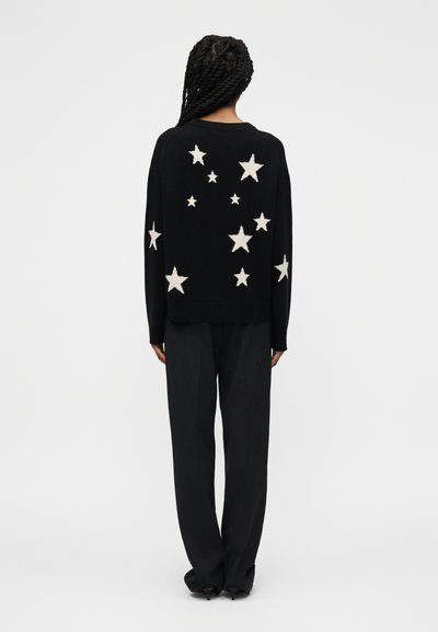 Άτομο με κοτσίδες που φοράει μαύρο πουλόβερ με λευκά αστέρια στην πλάτη και τα μανίκια, συνδυασμένο με άνετο μαύρο παντελόνι.