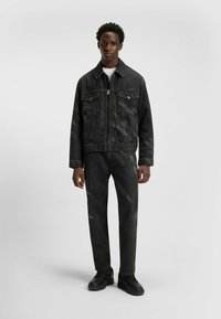 Zwarte spijkerjack met twee borstzakken, gecombineerd met bijpassende zwarte jeans met een subtiel patroon. De outfit wordt compleet gemaakt met zwarte schoenen.