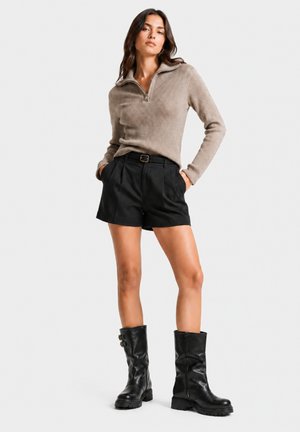 Donna con maglione beige a coste con cerniera, pantaloncini neri con cintura e stivali di pelle neri a metà polpaccio, in piedi con le mani in tasca su sfondo bianco.