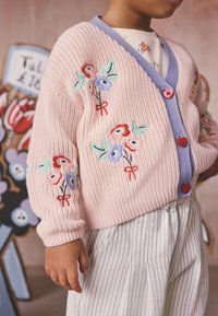 Child wearing a pink knit cardigan with floral embroidery and heart-shaped buttons over striped pants.