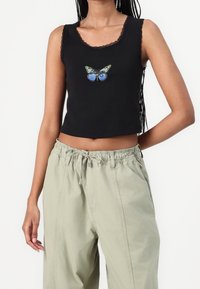 Top corto sin mangas negro con encaje y gráfico de mariposa combinado con pantalones verdes claros con cordón en una persona de pie.
