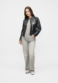 Γυναίκα που στέκεται με το χέρι στην τσέπη, φοράει μαύρο γυαλιστερό μπουφάν, μπεζ τοπ, ανοιχτογκρι φαρδιά τζιν παντελόνια και μπεζ παπούτσια σε λευκό φόντο.