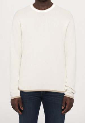 Mann trägt einen weißen, langärmeligen, strukturierten Pullover mit beigem Besatz und dunklen blauen Jeans, steht vor einem einfarbigen Hintergrund.