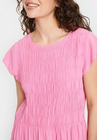 Robe rose avec une texture froncée, encolure arrondie et manches courtes. Le tissu tombe doucement, créant une silhouette décontractée.