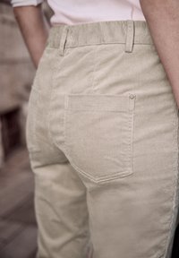 Personne portant un pantalon beige en velours côtelé avec une poche arrière visible et des passants de ceinture, debout à l'extérieur près d'un arrière-plan flou.
